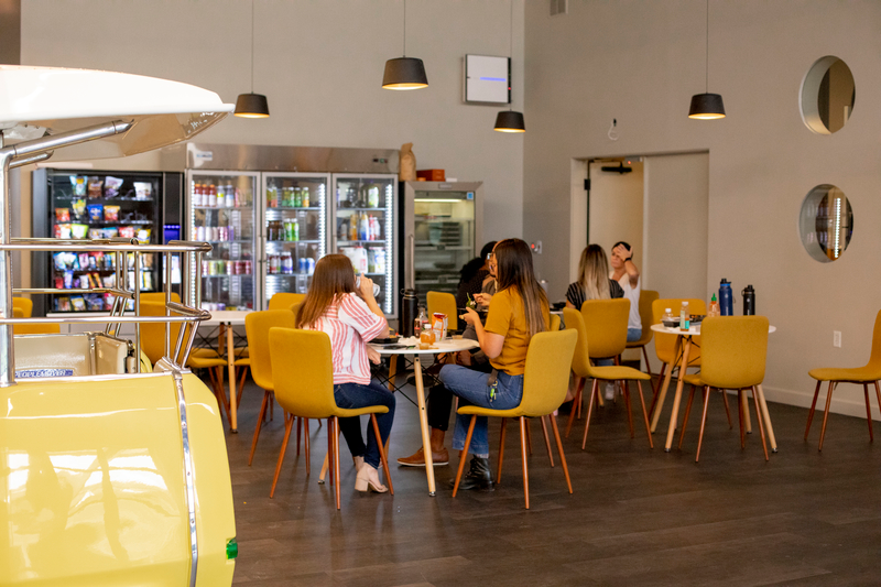 一群坐着的员工在公司用餐区愉快地社交，用餐区摆满了小桌子和黄色椅子，前面是一排自动贩卖机和储备充足的饮料机